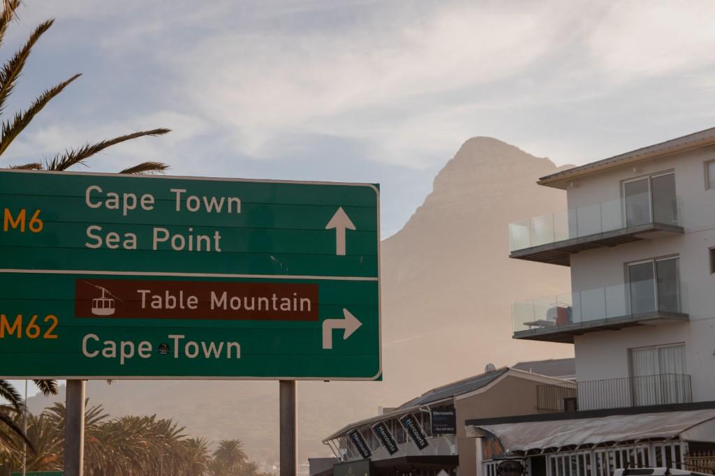 Cape Town road sign for M6 and M62 toward Sea Point and Table Mountain cable car, with Lion's Head and coastal apartment buildings in the background.