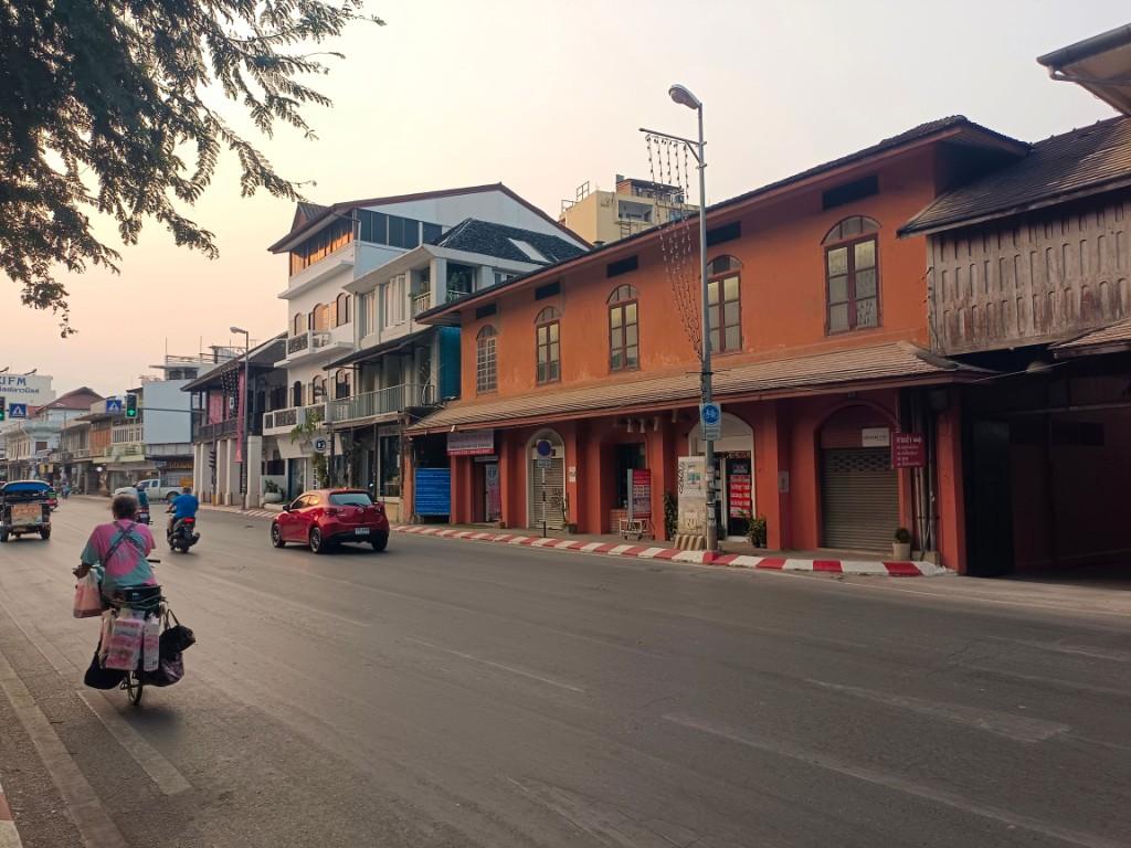 Chiang Mai street at weekday pace with a cyclist carrying goods, cars, a motorcycle and terracotta shopfronts under soft morning light.